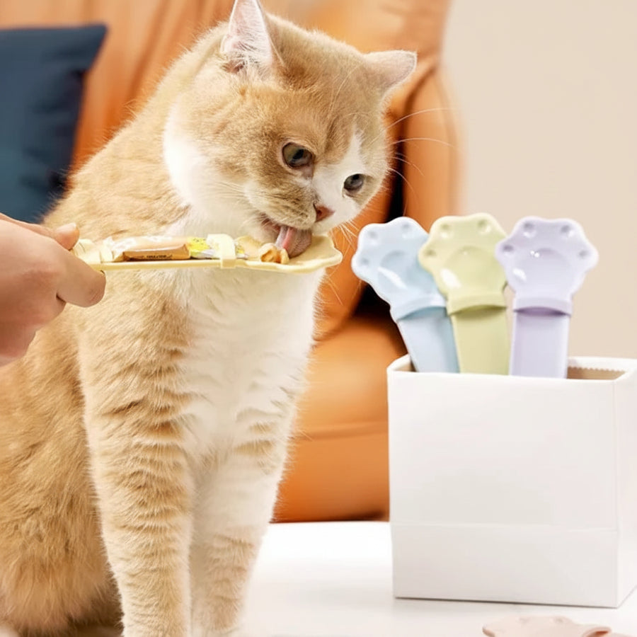 An orange tabby cat licks a small plate held by a human hand, with a white container of colorful cat treats or lick mats nearby. The scene is set in a cozy indoor environment with an orange couch in the background.